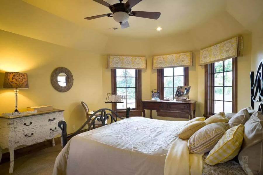 Bedroom furnished with an ornate iron bed, a distressed dresser, and a dark wood desk against the bay window.