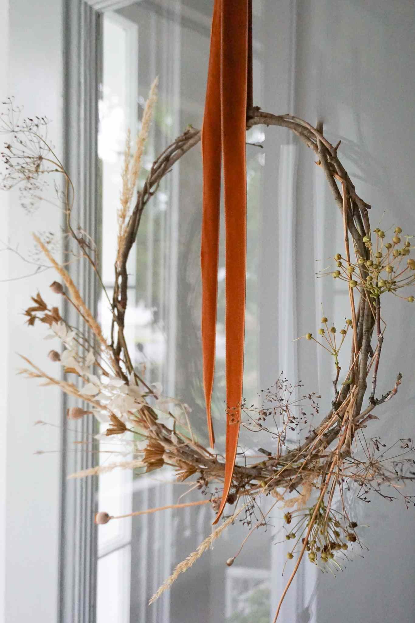 Airy foraged wreath of dried grasses and twigs hanging in a window from a long orange velvet ribbon