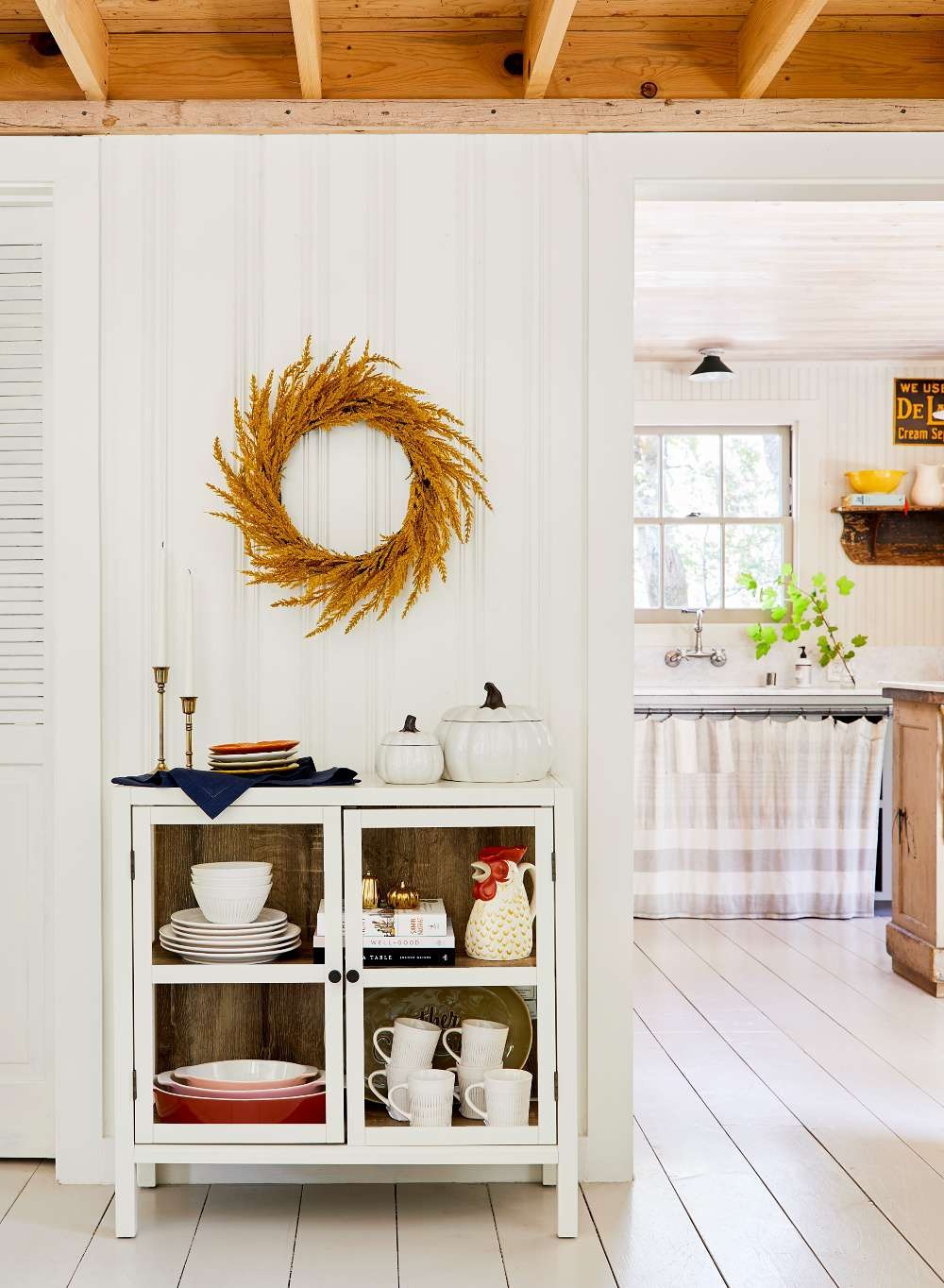 Golden dried-grass wreath hung above a white glass-front cabinet in a bright farmhouse kitchen