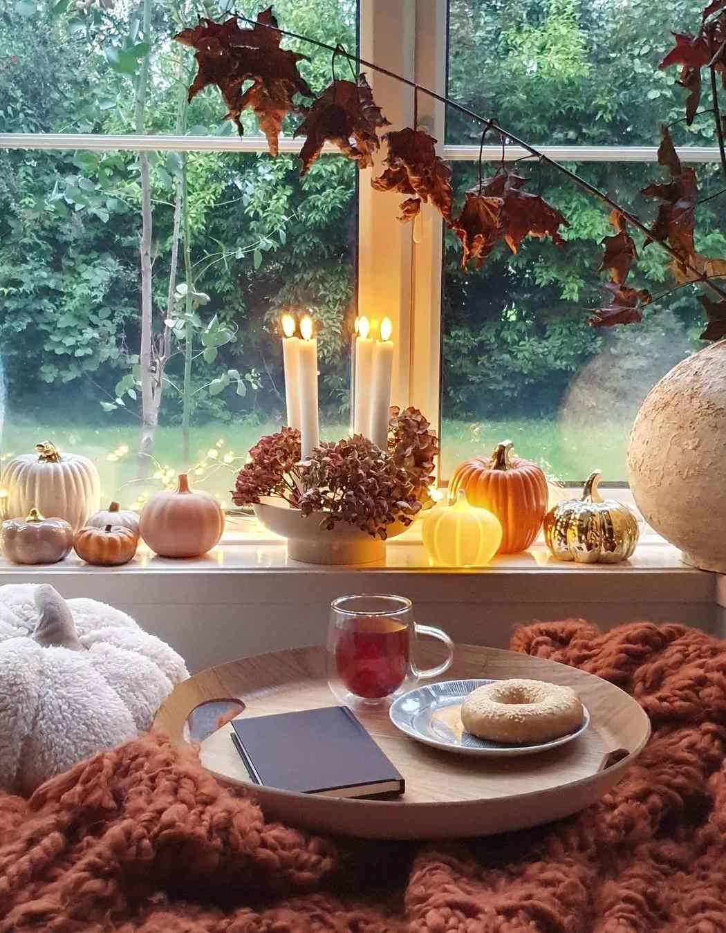 Cozy fall window nook with faux pumpkins, lit taper candles, dried hydrangeas and autumn leaves above a tray with tea